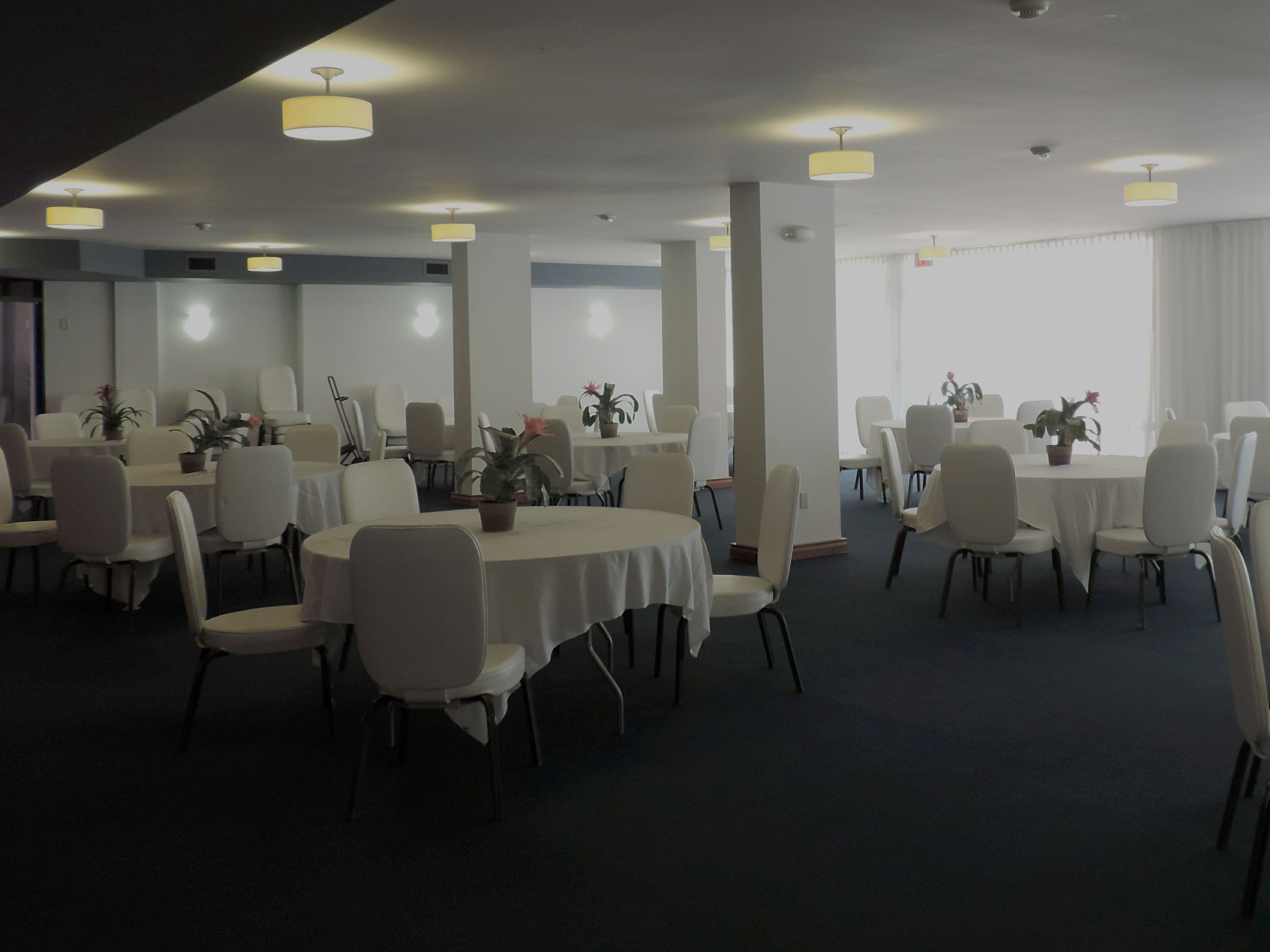 A spacious conference room with multiple tables and chairs, large windows with curtains, and ceiling lights.