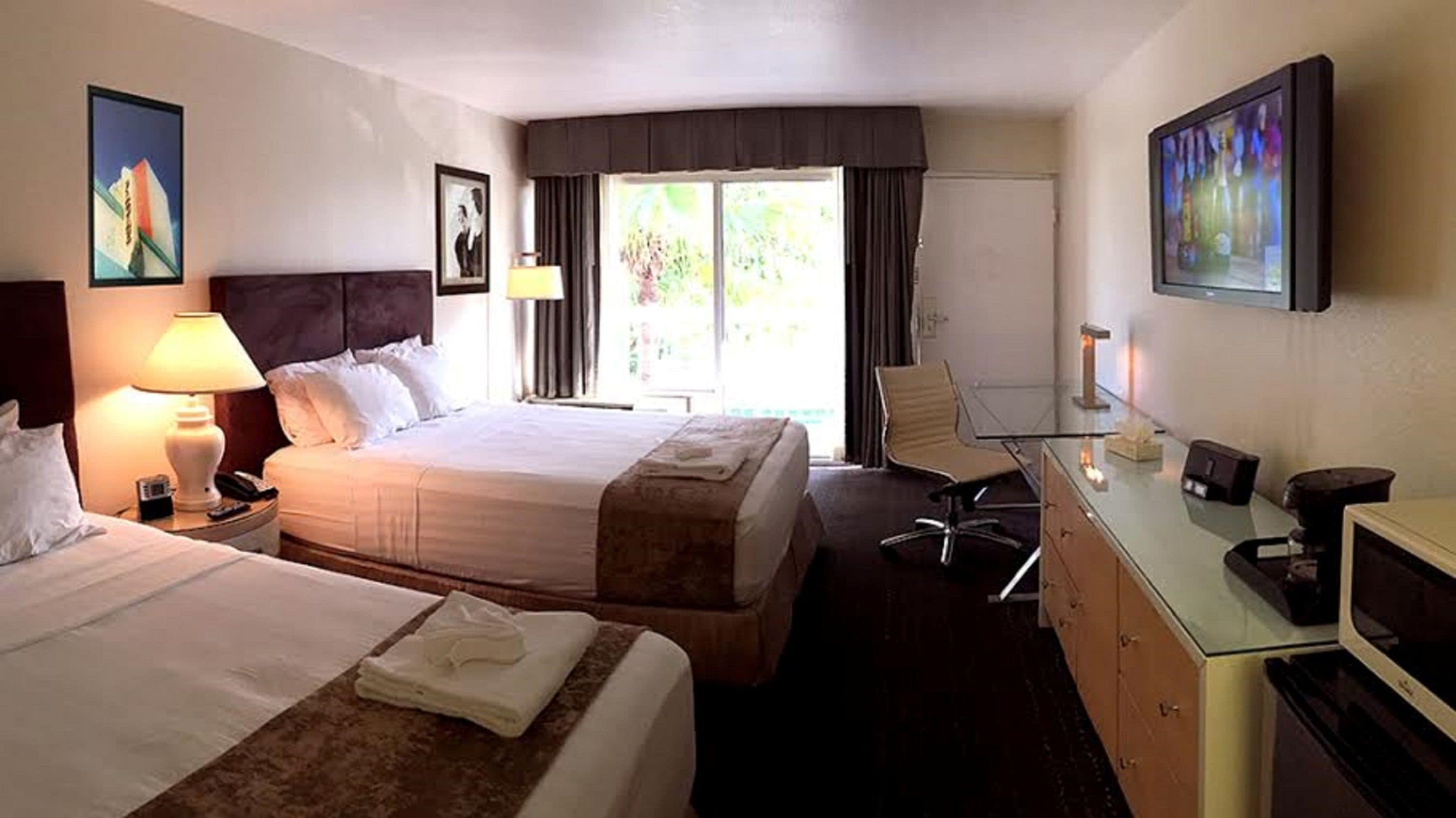 Ein Hotelzimmer mit zwei Betten, einem Schreibtisch mit Computer, einem Sessel, einer Lampe und Kunstwerken an den Wänden.