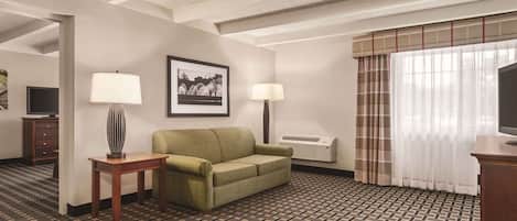 A hotel room with a patterned carpet, a green sofa, two lamps, a framed picture on the wall, and a television on a wooden stand.