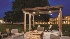 Een terras buiten met een vuurkorf van steen, een houten pergola en zitplaatsen.