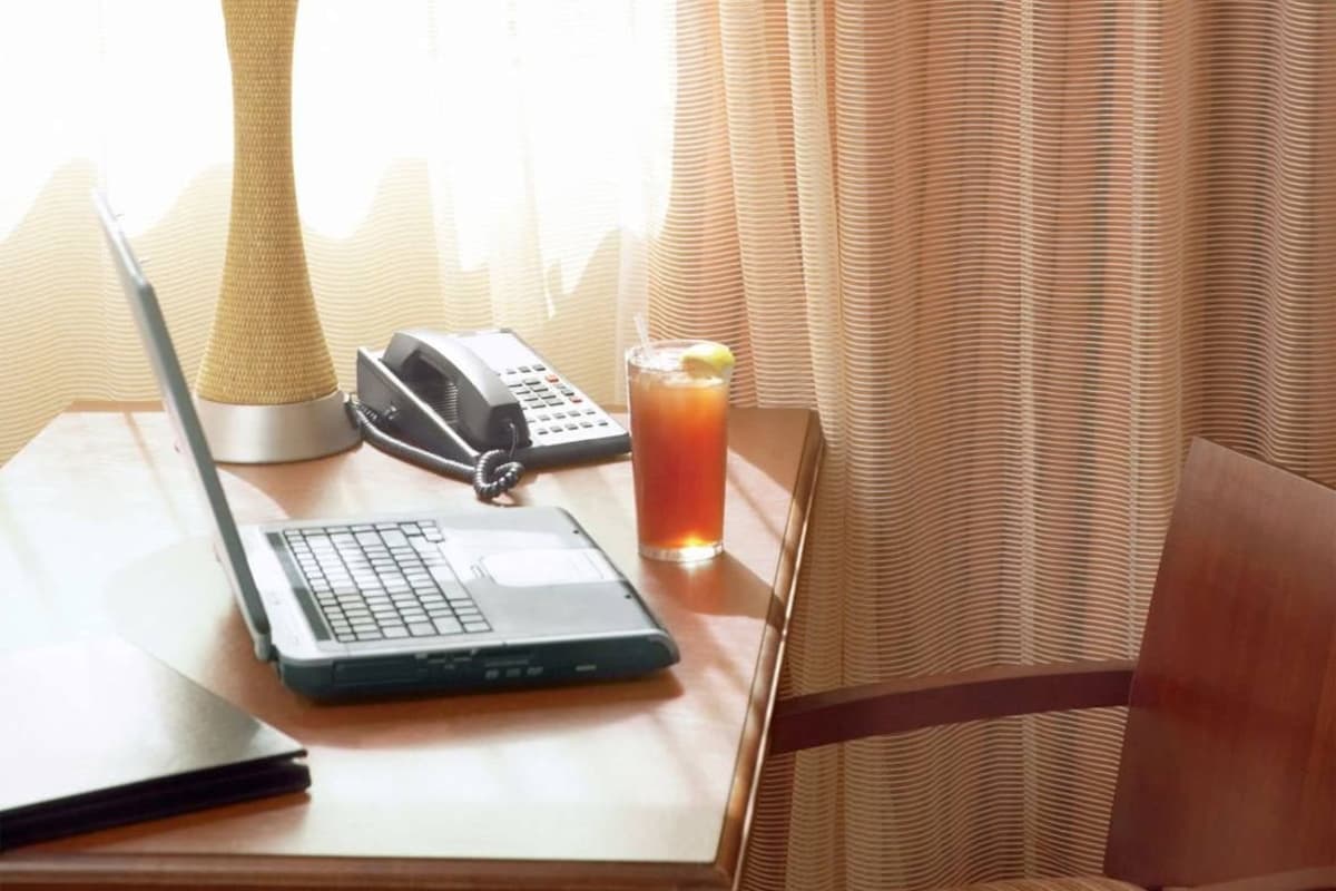 A hotel room with a laptop on a table, a telephone, a glass of iced tea, a lamp, and a chair.
