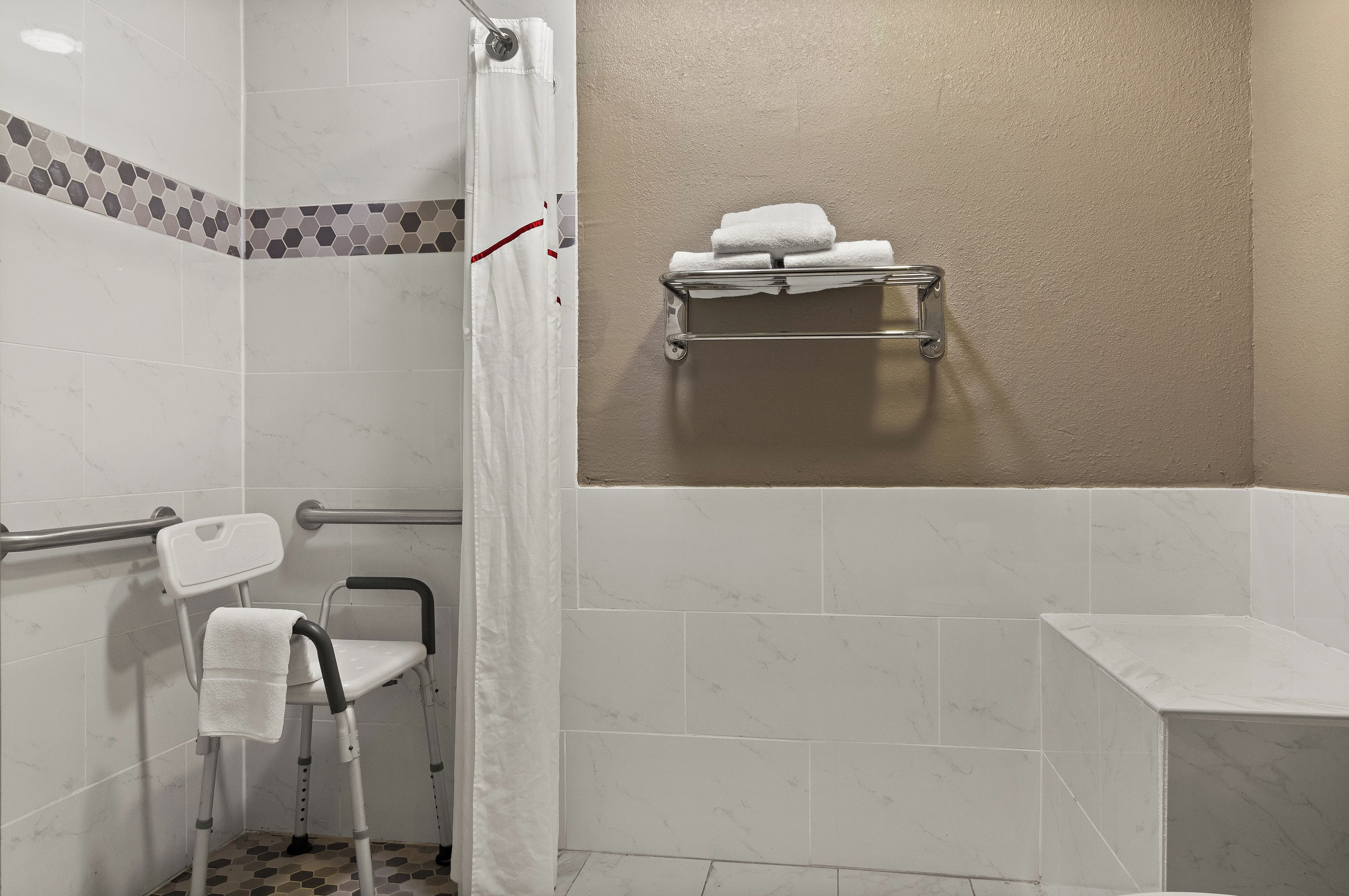 A bathroom with a shower, a grab bar, a wheelchair, a towel rack, and a white tiled floor.