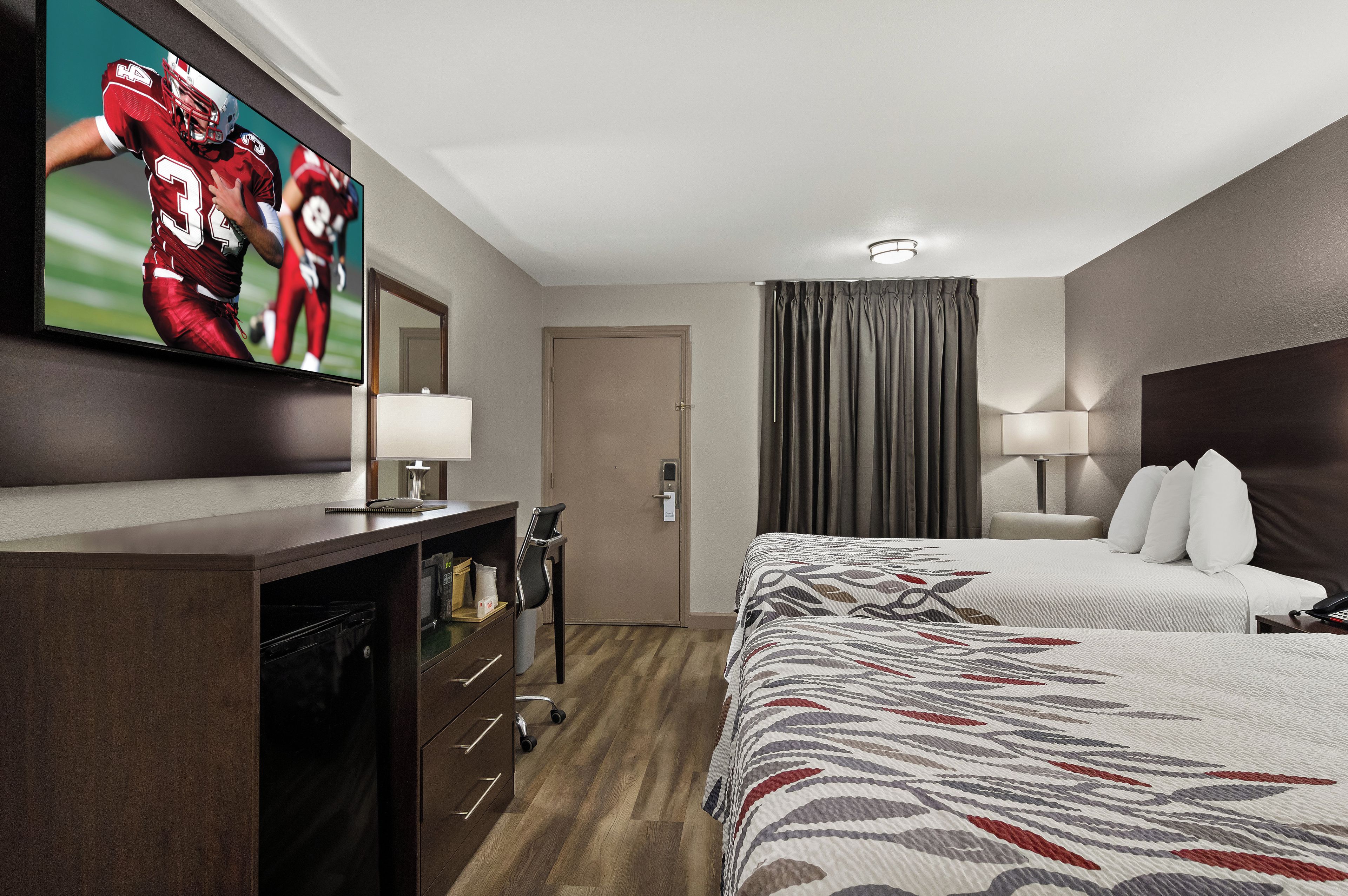 A hotel room with a large flat-screen TV on the wall, a bed with a patterned bedspread, a desk with a lamp, and a door leading to the bathroom.
