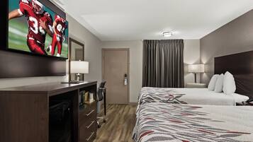 A hotel room with a large flat-screen TV on the wall, a bed with a patterned bedspread, a desk with a lamp, and a door leading to the bathroom.