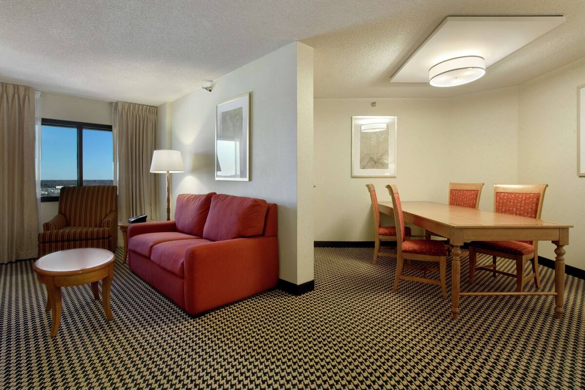 A hotel room with a red sofa, a small round table, a dining table with four chairs, a lamp, and framed artwork on the walls.