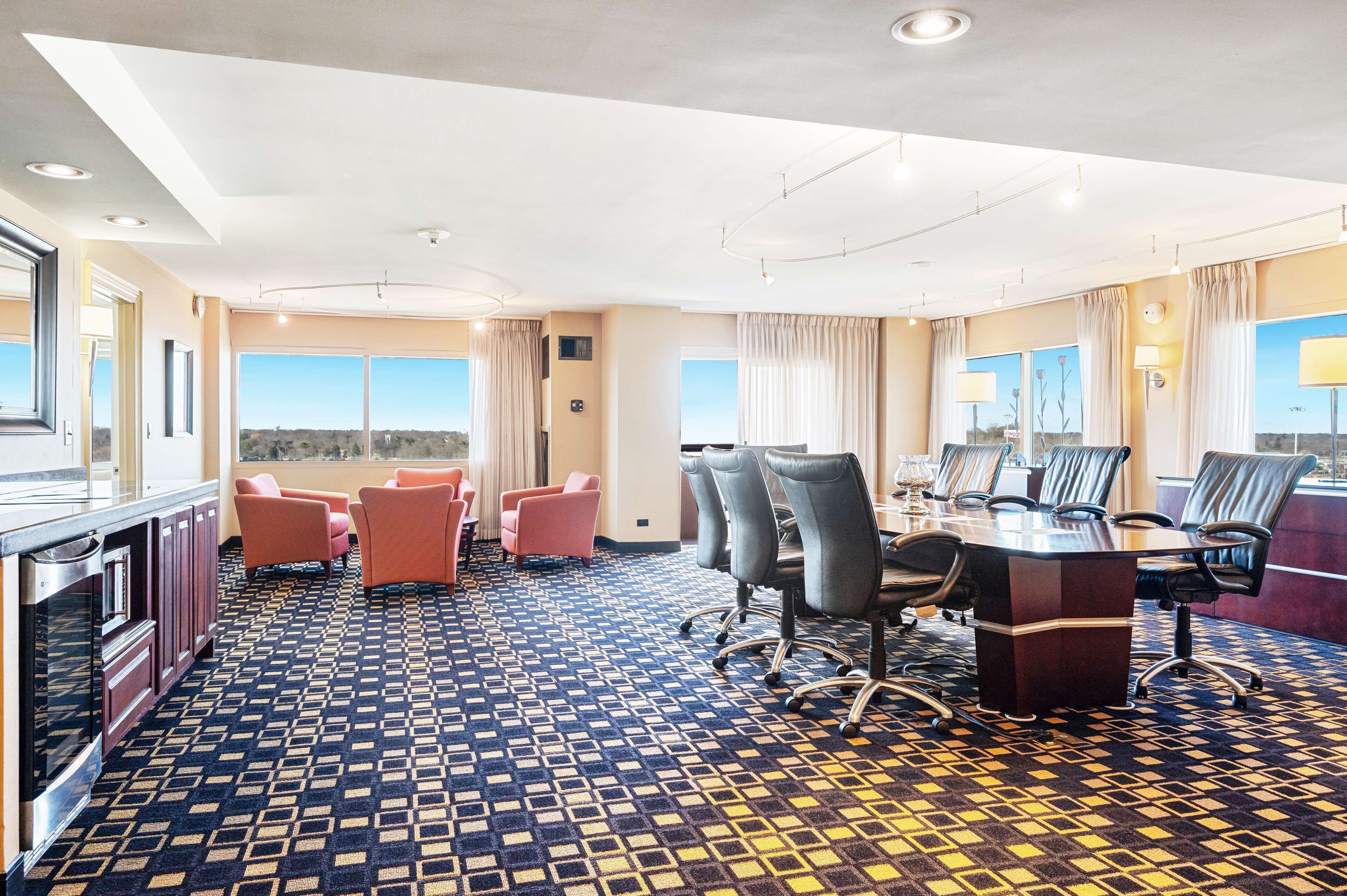 Un salón de conferencias con una gran mesa y varias sillas, una alfombra estampada y ventanas grandes con cortinas.