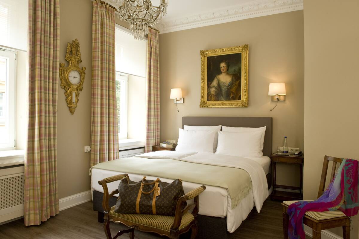 A hotel room with a large bed, two bedside tables, a chandelier, a painting on the wall, and a window with curtains.