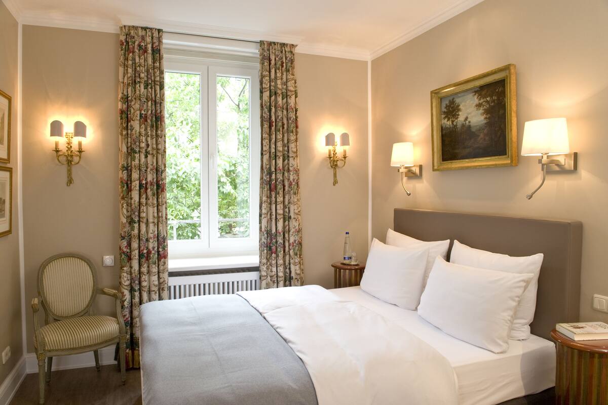 A hotel room with a bed, a chair, a window with curtains, wall-mounted lamps, and a painting.