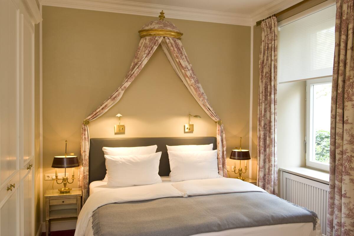 A neatly made bed with a canopy, two bedside lamps, and a window with curtains.