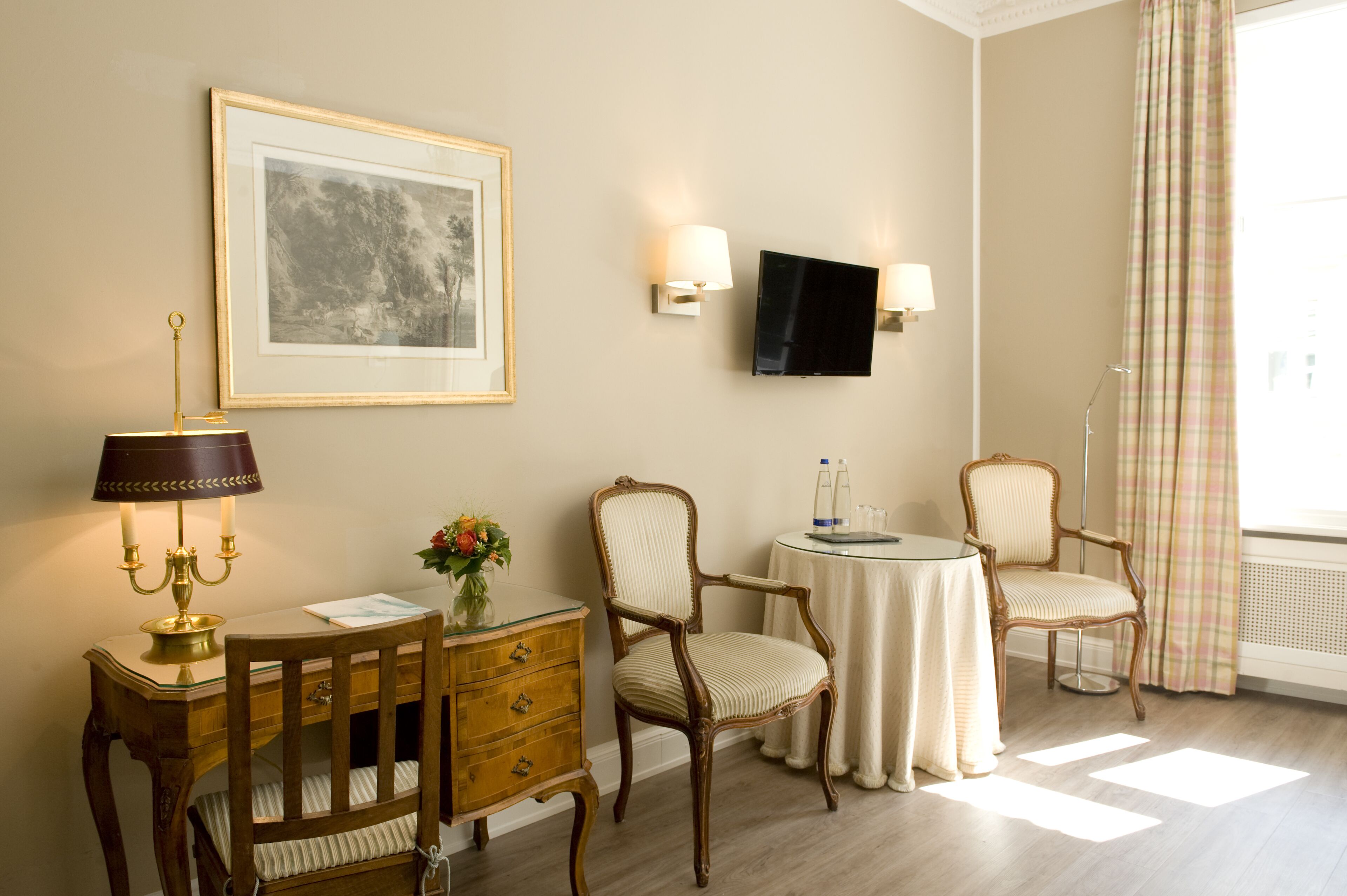 Une pièce comprenant un bureau en bois, deux chaises, une petite table, un tableau encadré, des lampes fixées au mur et un téléviseur.