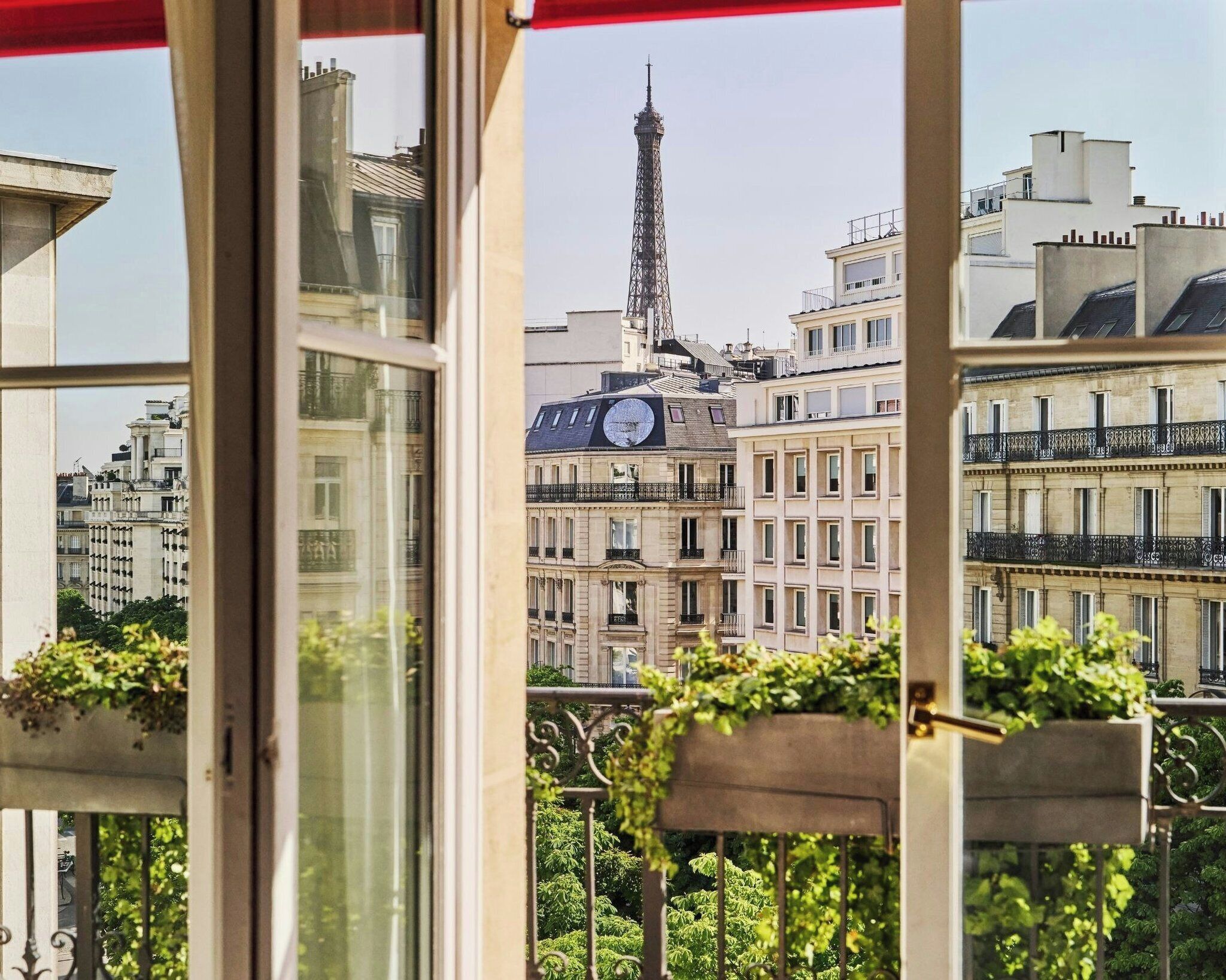 Vista a través de una ventana con balcón, mostrando un edificio con un reloj y la Torre Eiffel al fondo.