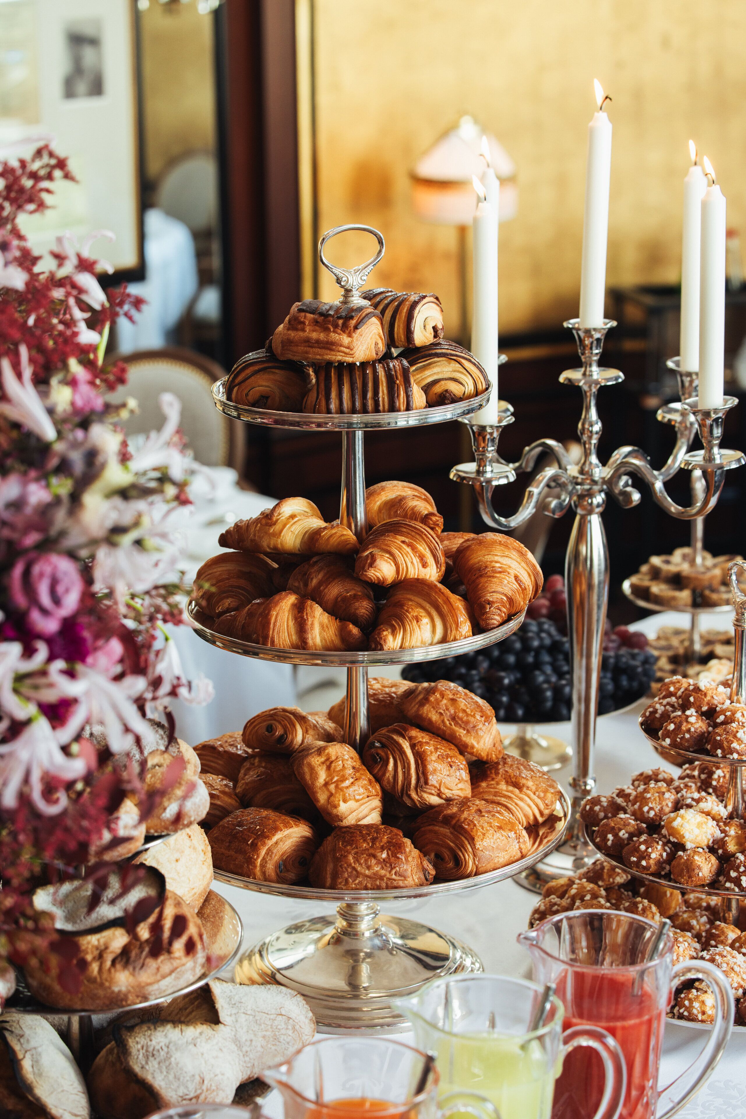 Une présentation à plusieurs niveaux de viennoiseries et de pains, avec un chandelier et une composition florale en arrière-plan.