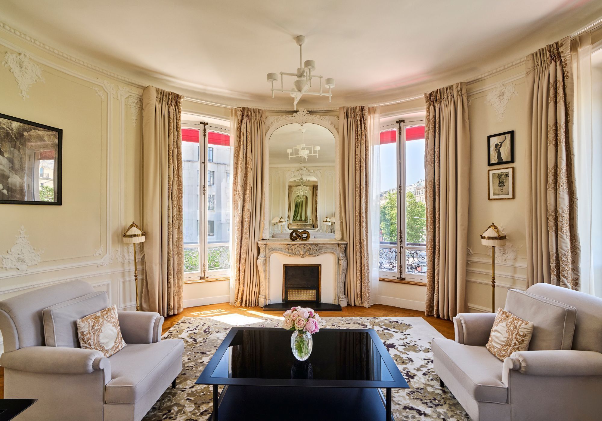 Una habitación con chimenea, dos sofás, una mesa de centro con flores y ventanas grandes con cortinas.