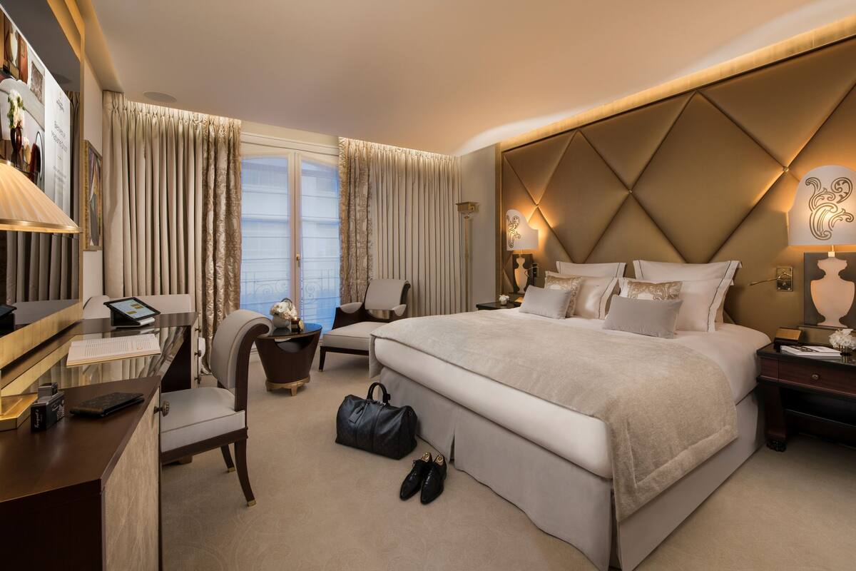 A hotel room with a large bed, a desk, a chair, and a nightstand with a lamp.