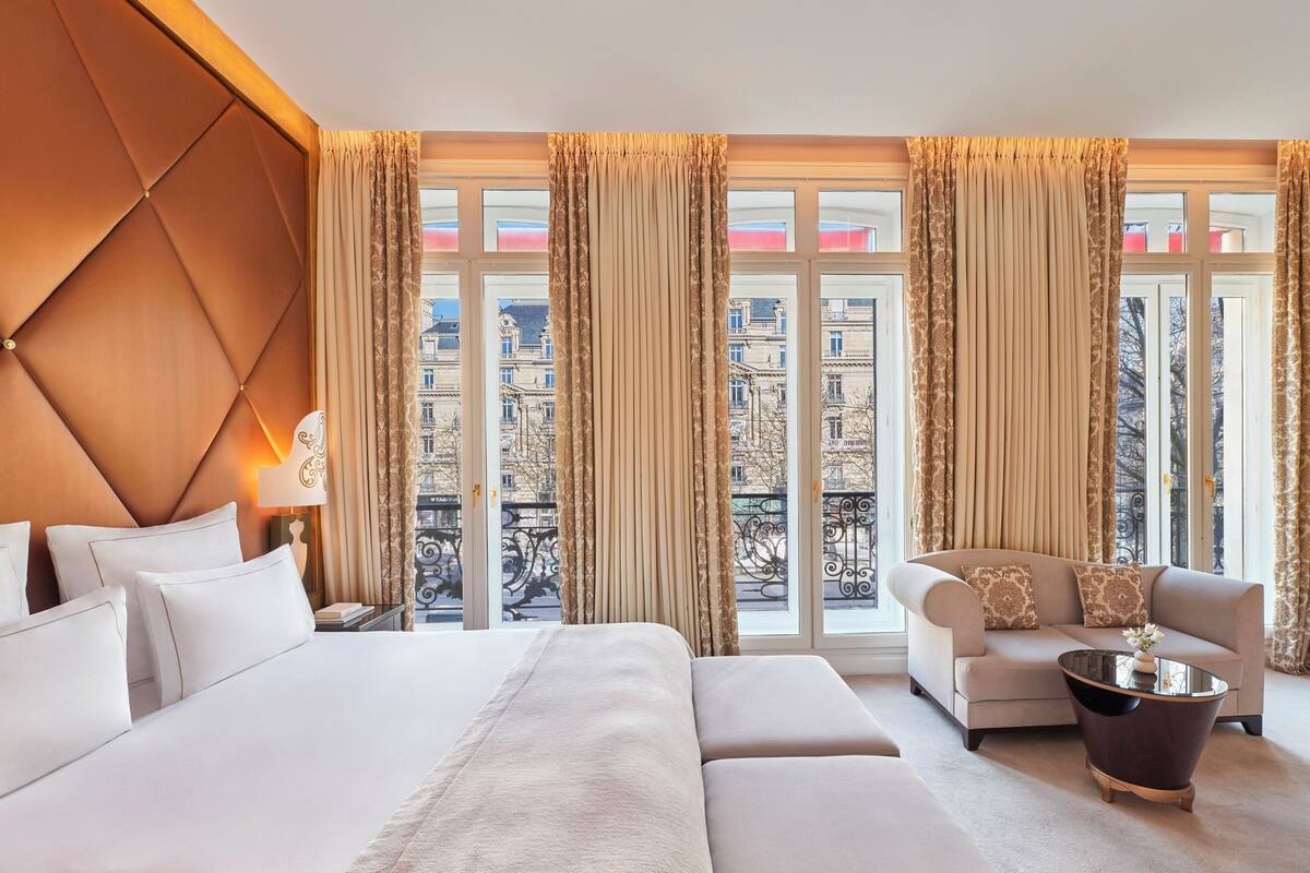 A hotel room with a large bed, a sofa, a coffee table, and a view of the city through large windows.