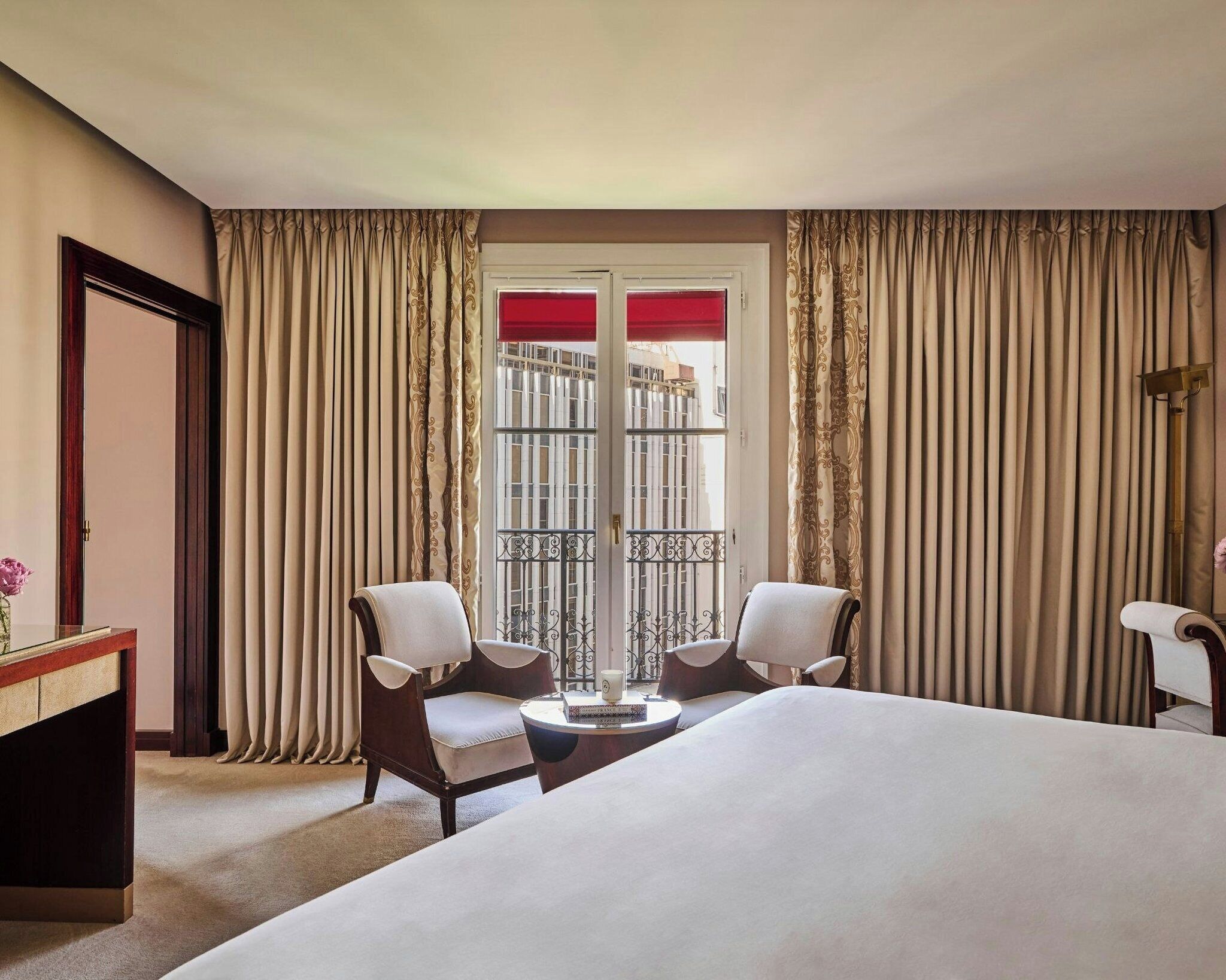 A hotel room with a bed, two armchairs, a small table, and a view of the outside through a window with curtains.