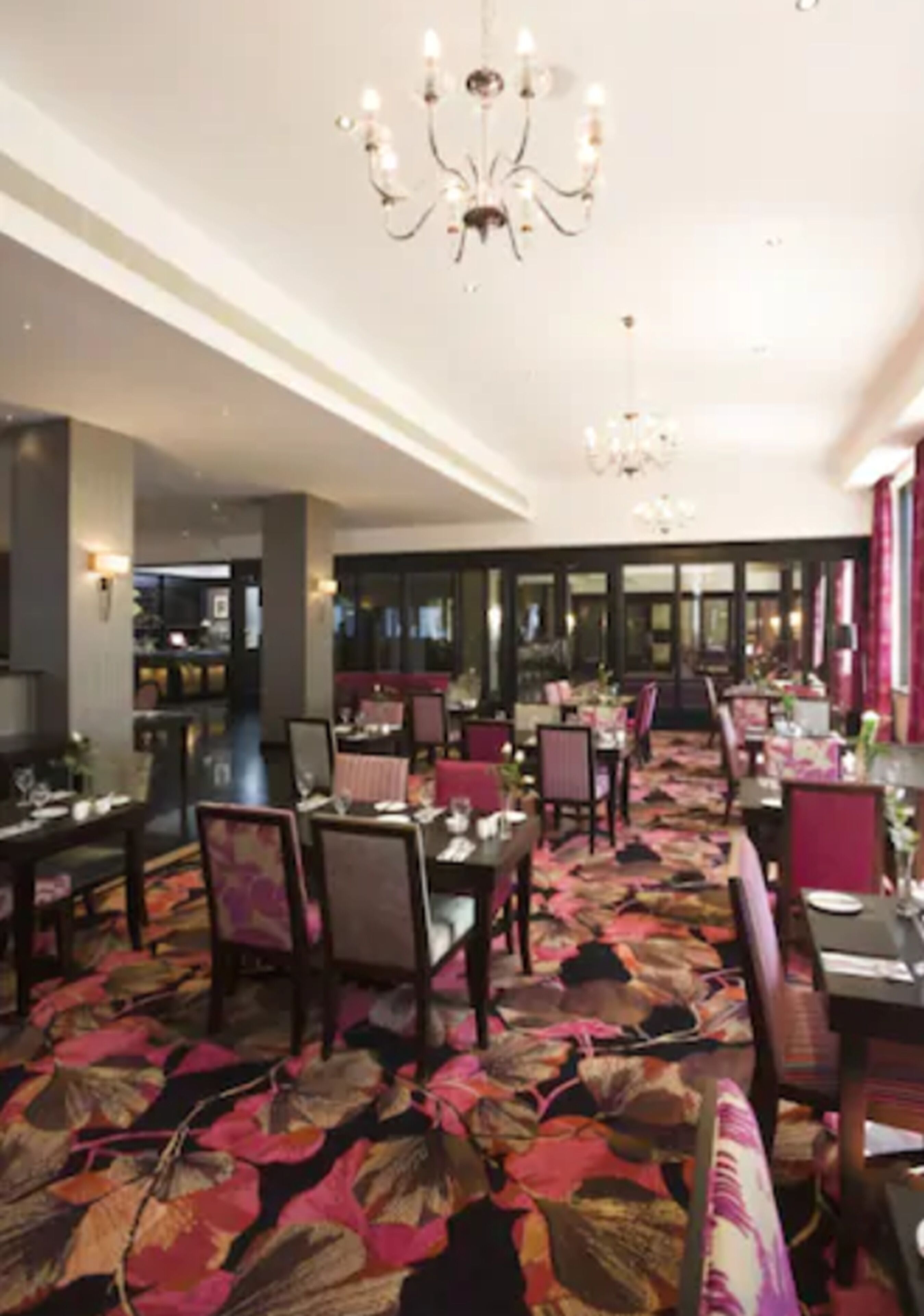 A hotel dining area with tables set for guests, a patterned carpet, and chandeliers.