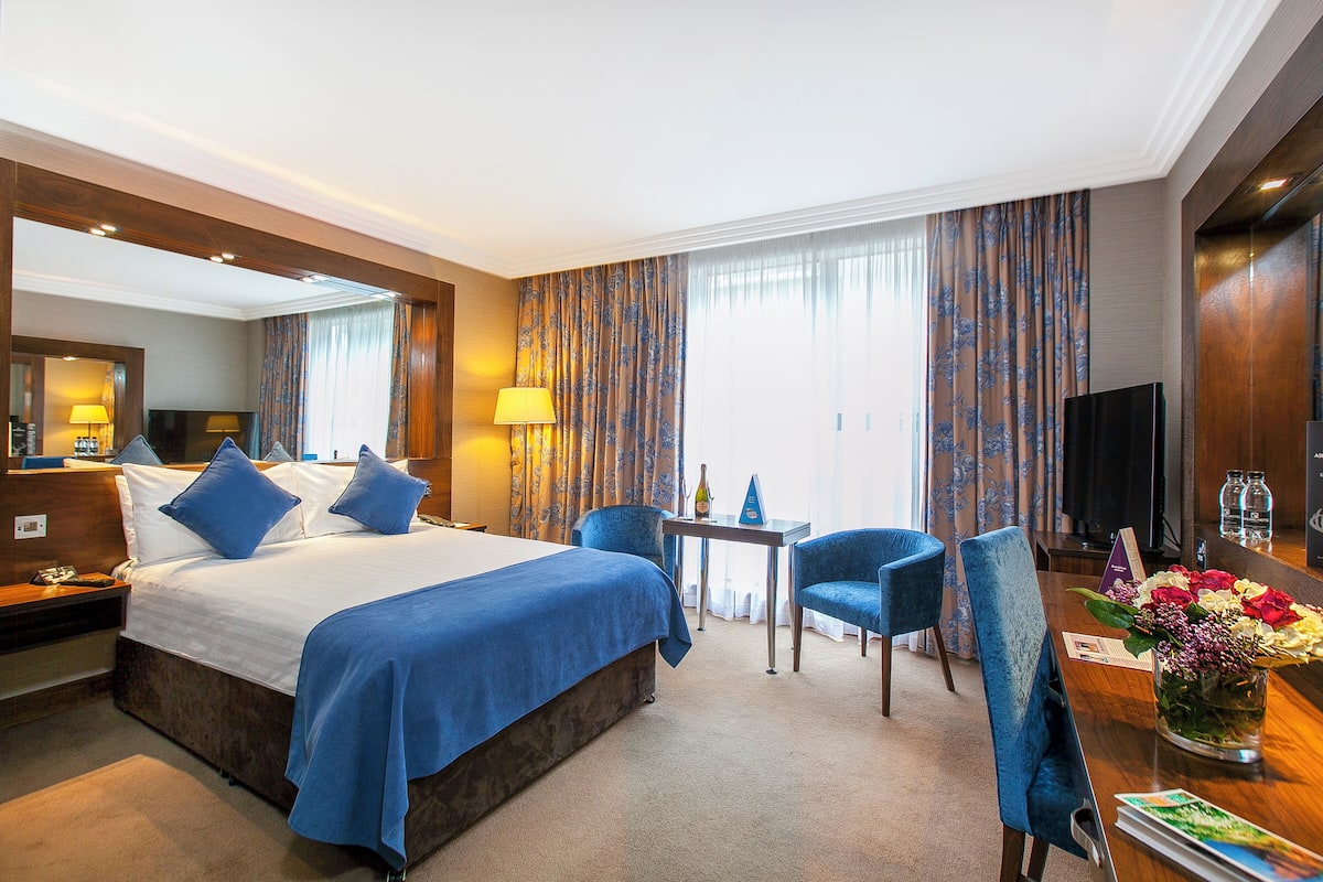 Habitación de hotel con una cama grande, dos sillas azules, una mesita, un televisor y un florero con flores.