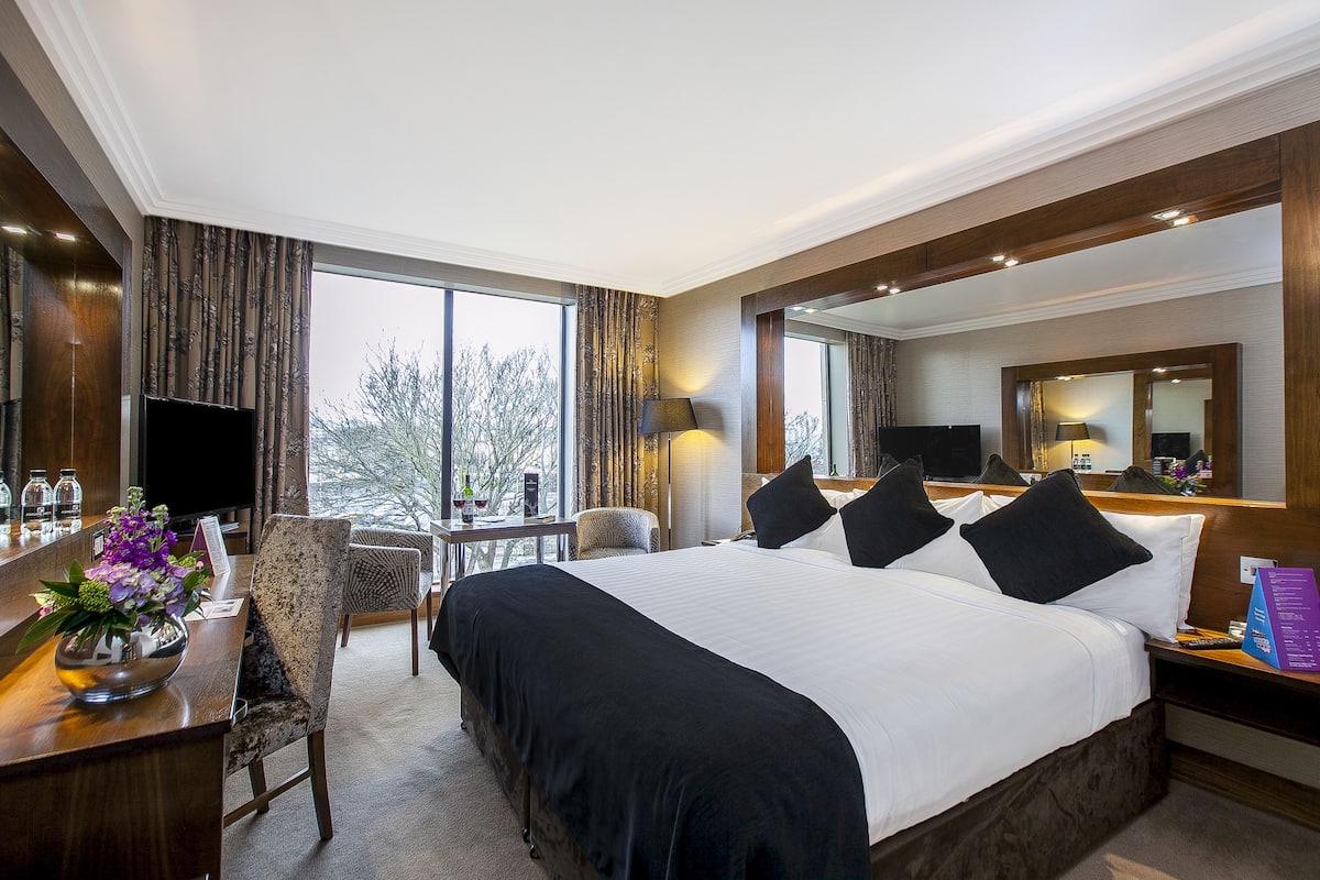 Una habitación de hotel con una cama grande, un escritorio con computadora, un televisor y una ventana con vista a árboles.