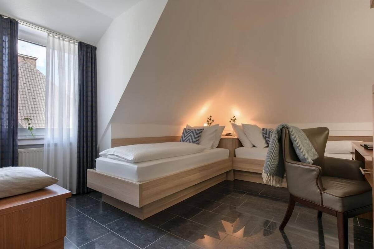 A modern hotel room with a bed, a chair, a nightstand, and a window with curtains.