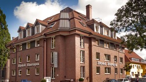 Un immeuble en briques de plusieurs étages, portant l’enseigne « Hôtel Europa », avec une entrée imposante, de grandes fenêtres et un espace de stationnement où sont garées des voitures.