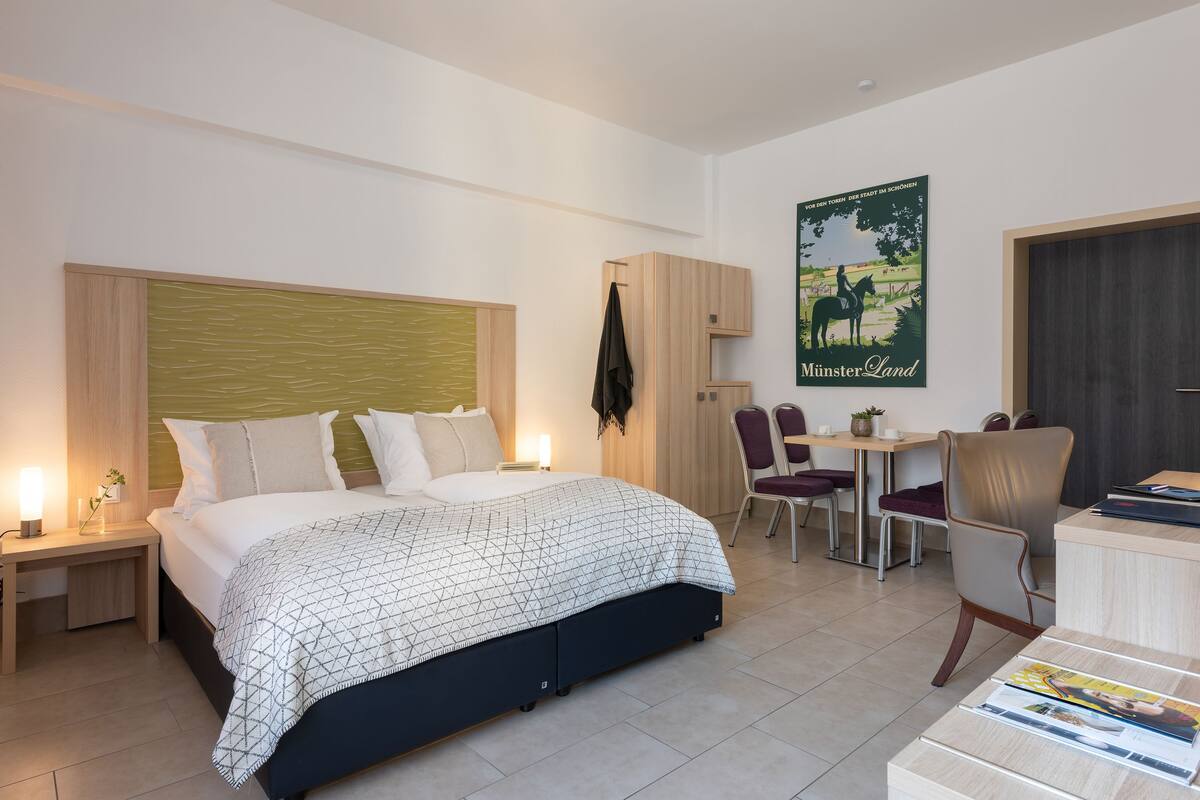 A modern hotel room with a large bed, a desk, and a chair. There is a poster on the wall and a coat rack.