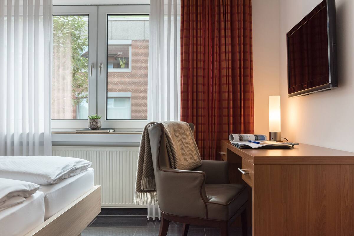 A hotel room with a bed, a desk with a lamp, a chair, and a window with curtains.