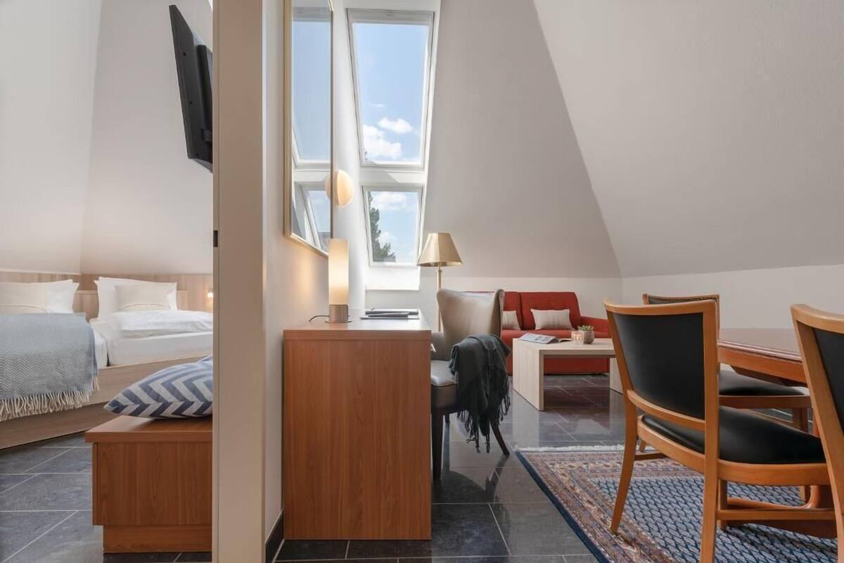 A modern hotel room with a bed, a desk, a chair, and a sofa. There are windows allowing natural light and a view of the outdoors.