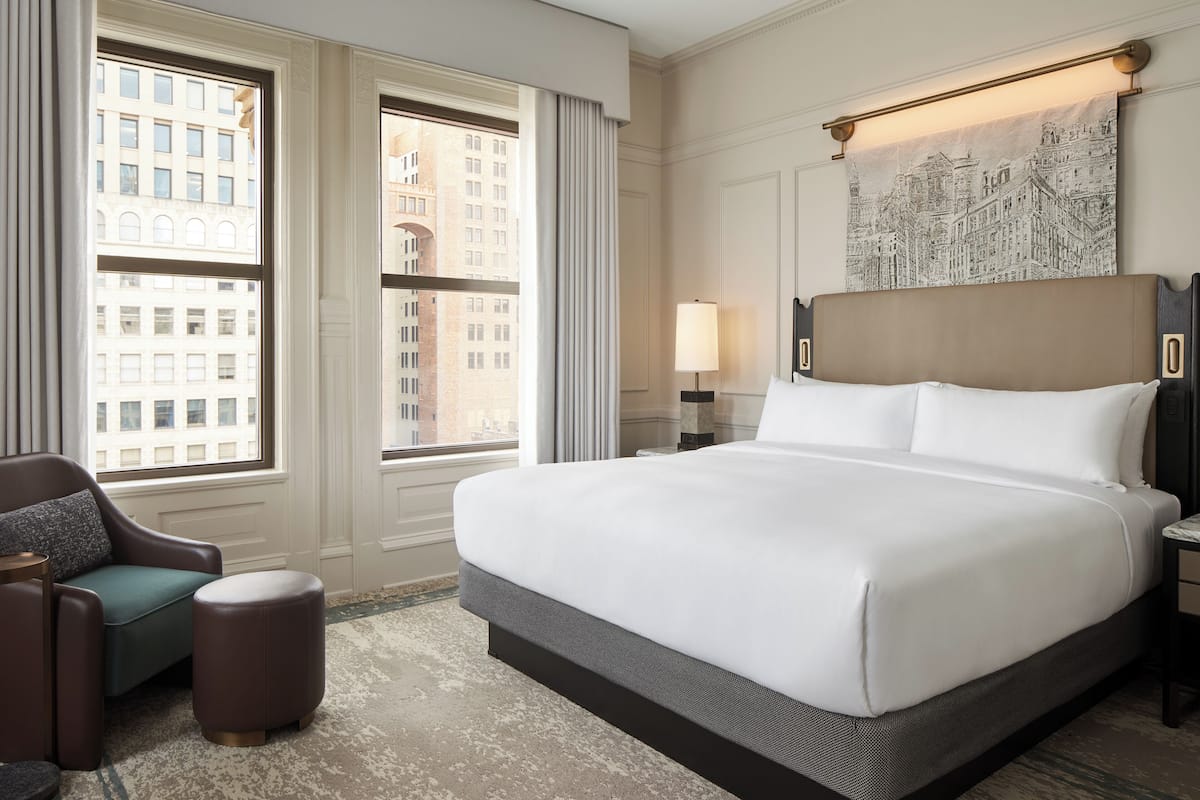 A hotel room with a large bed, a chair, a small ottoman, a nightstand with a lamp, and a cityscape view through the windows.