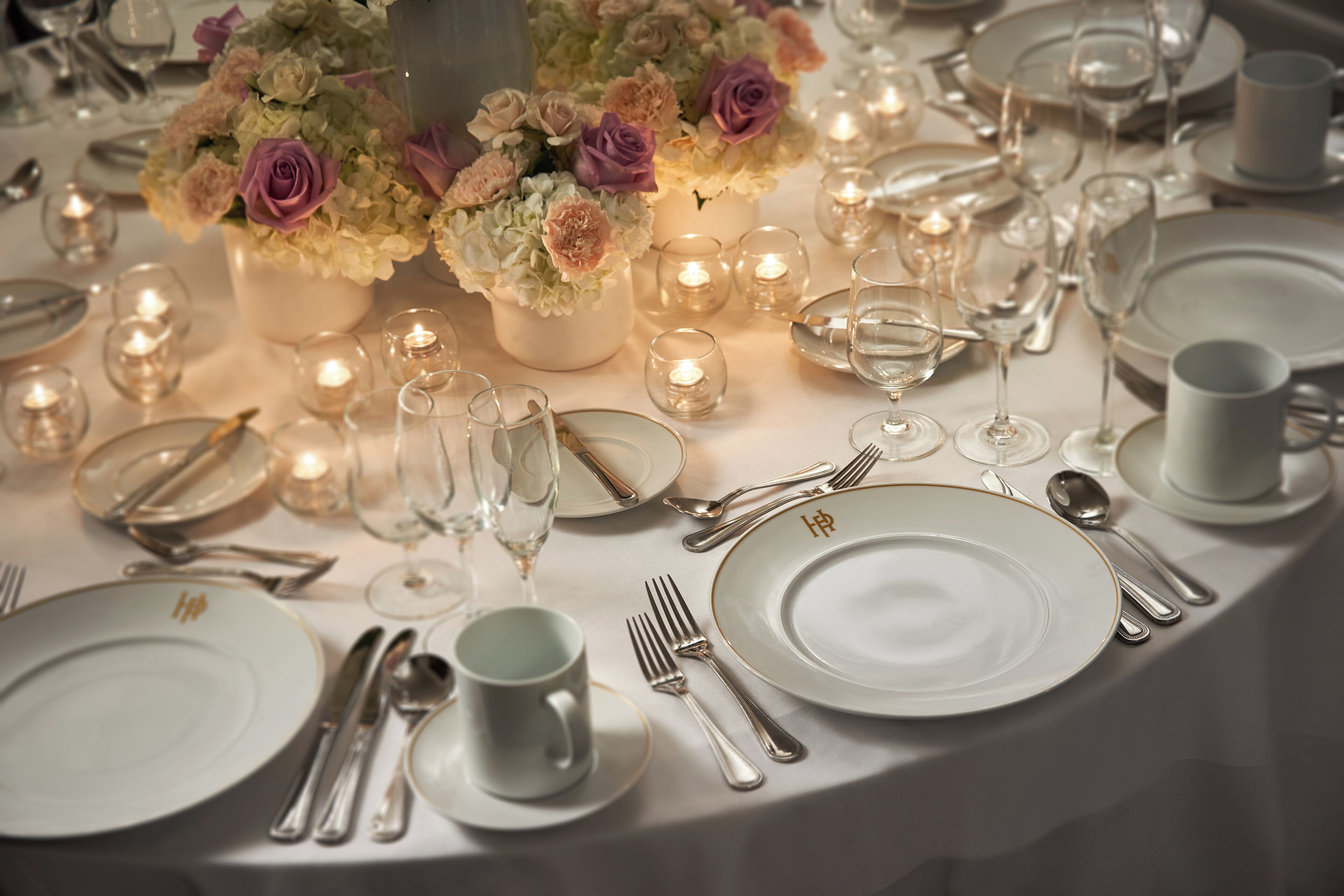 A table set for a formal dinner with white plates, silverware, and glassware, accompanied by small candles and floral centerpieces.