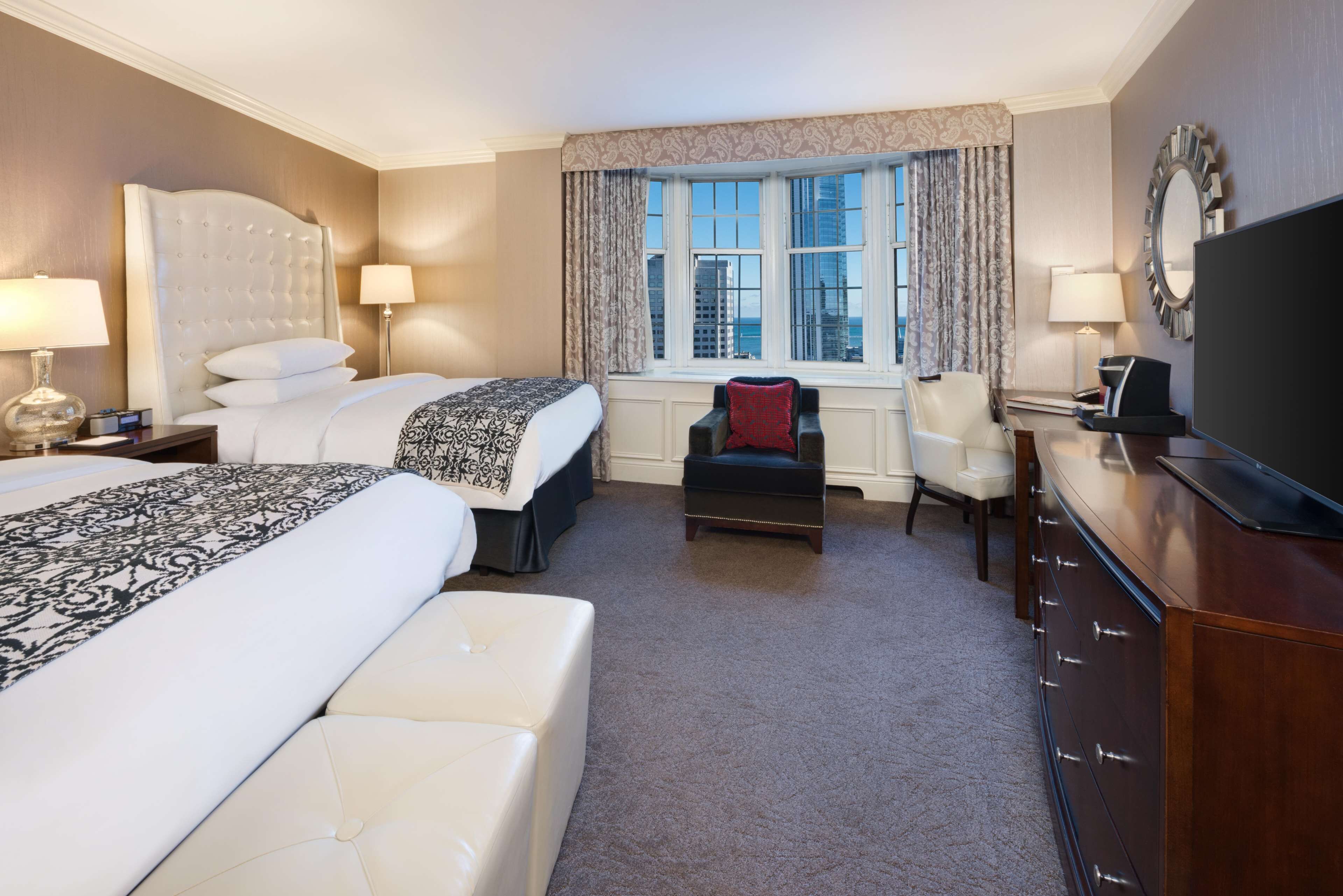 A hotel room with two beds, a desk, a chair, a TV, and a window with a view of buildings.