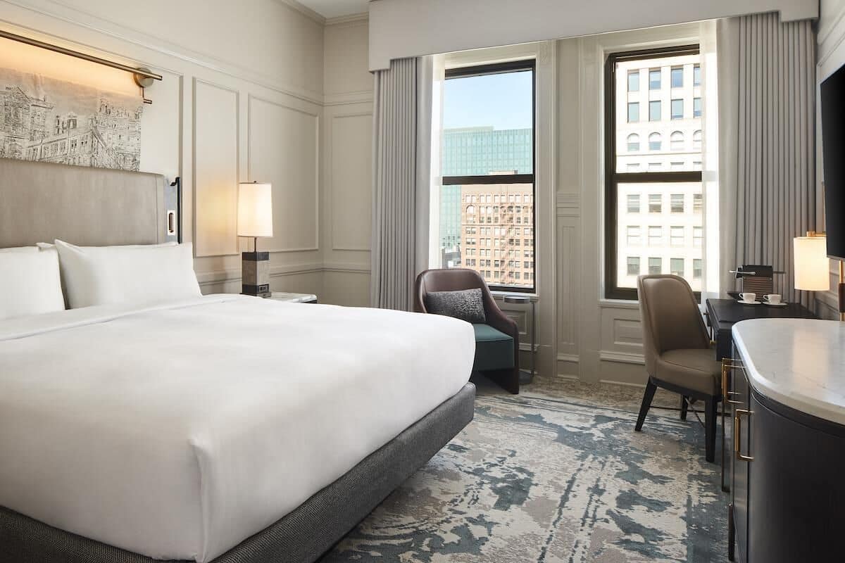 A hotel room with a large bed, a desk, a chair, and a view of the city through the windows.