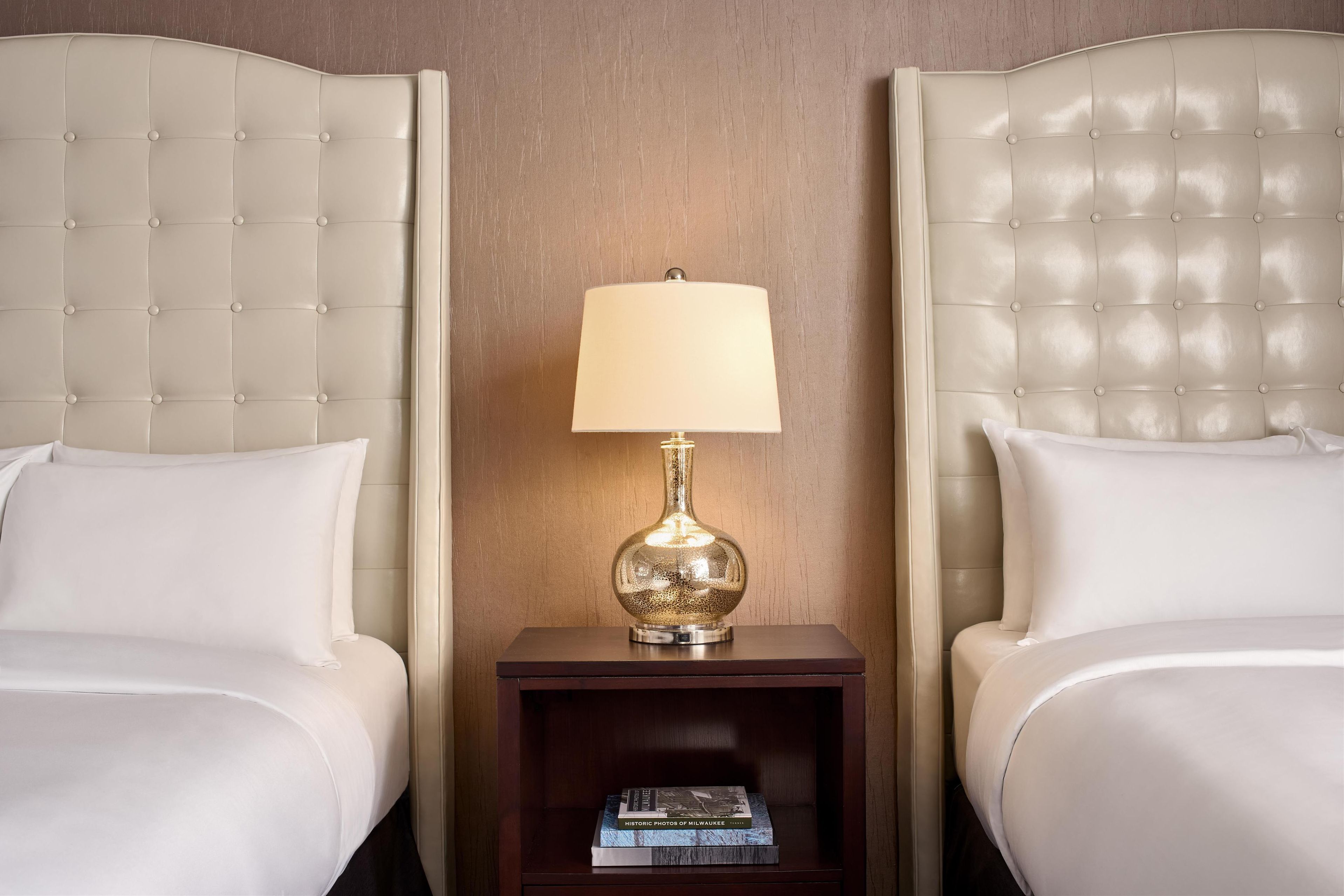A hotel room with two beds, a nightstand with a lamp, and a book on the nightstand.