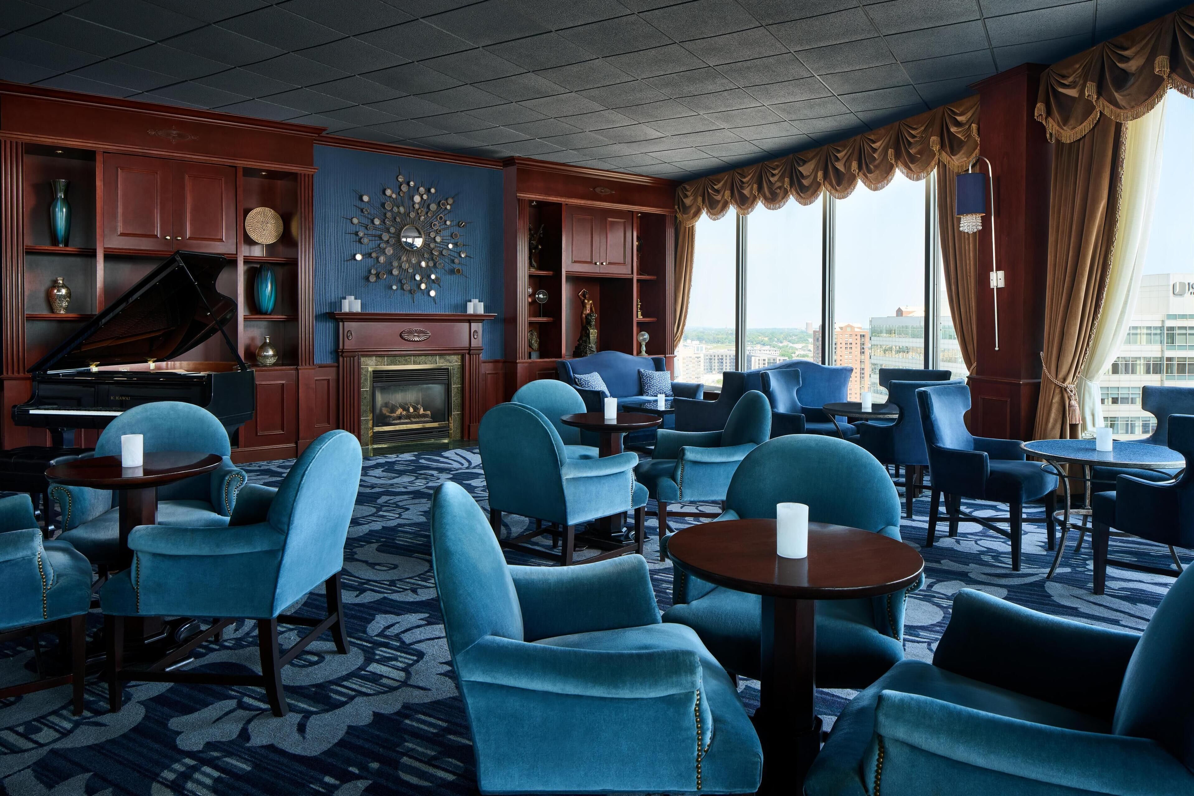 A hotel lobby with a grand piano, blue armchairs, a fireplace, and a city view.