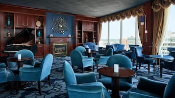 A hotel lobby with a grand piano, blue armchairs, a fireplace, and a city view.