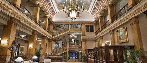 A grand lobby with ornate columns, a chandelier, and a staircase leading to an upper level.