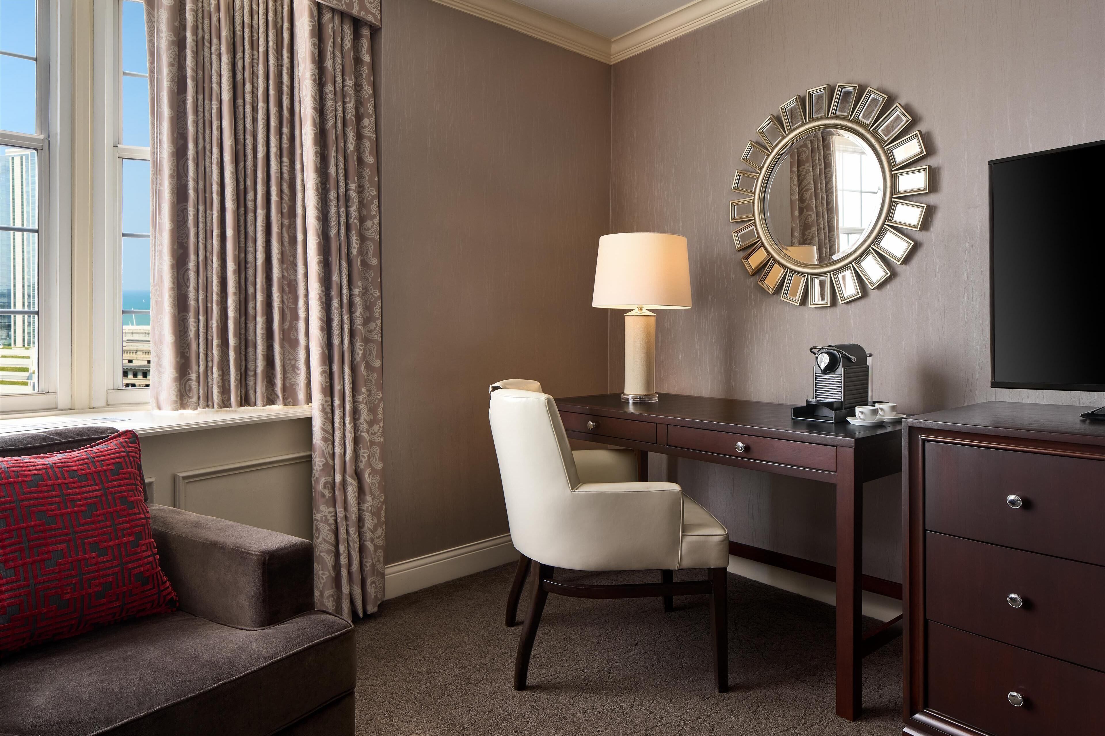 Une chambre d’hôtel avec une grande fenêtre, un bureau avec une lampe, une chaise, un téléviseur et une commode.