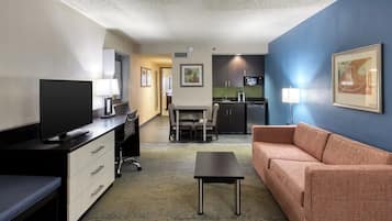 Een hotelkamer met televisie, bureau, stoel, bank, kitchenette en kunstwerken aan de wanden.