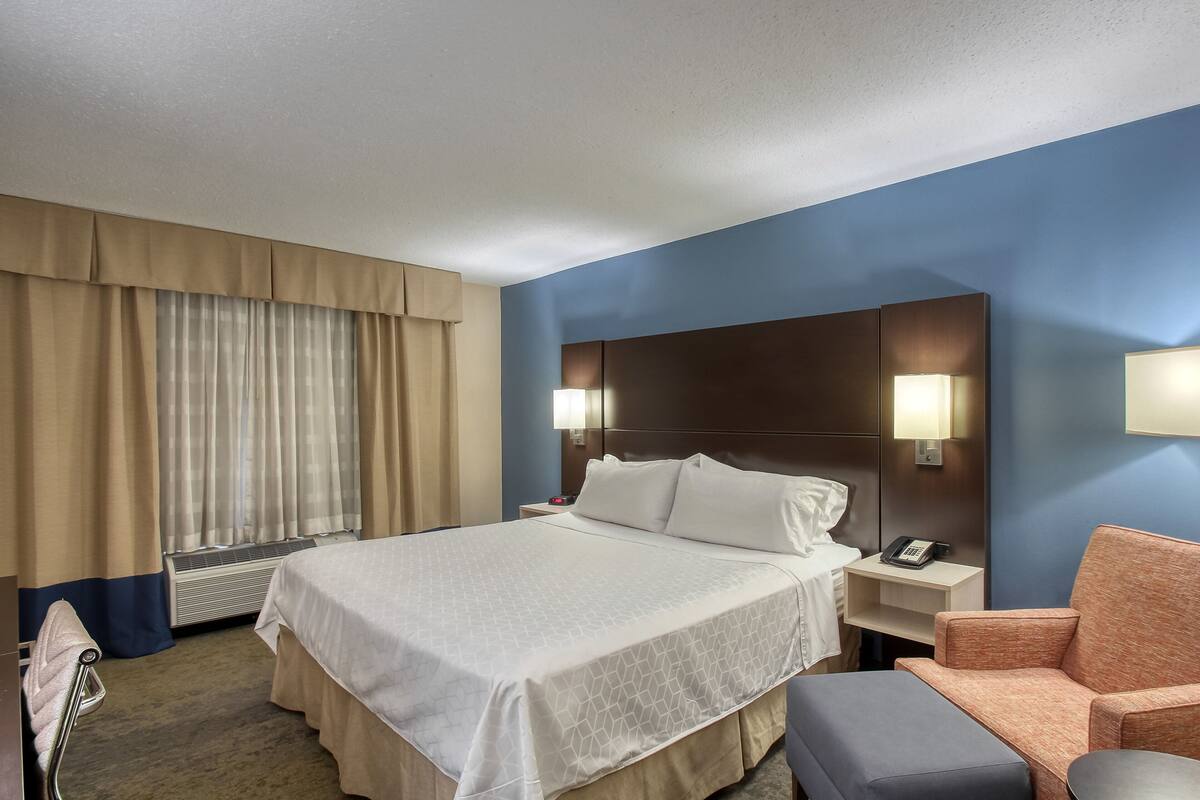 A hotel room with a large bed, a nightstand with a lamp, a chair, and a small table.