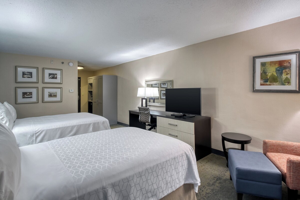 A hotel room with two beds, a desk with a chair, a television, a small round table, and a framed picture on the wall.