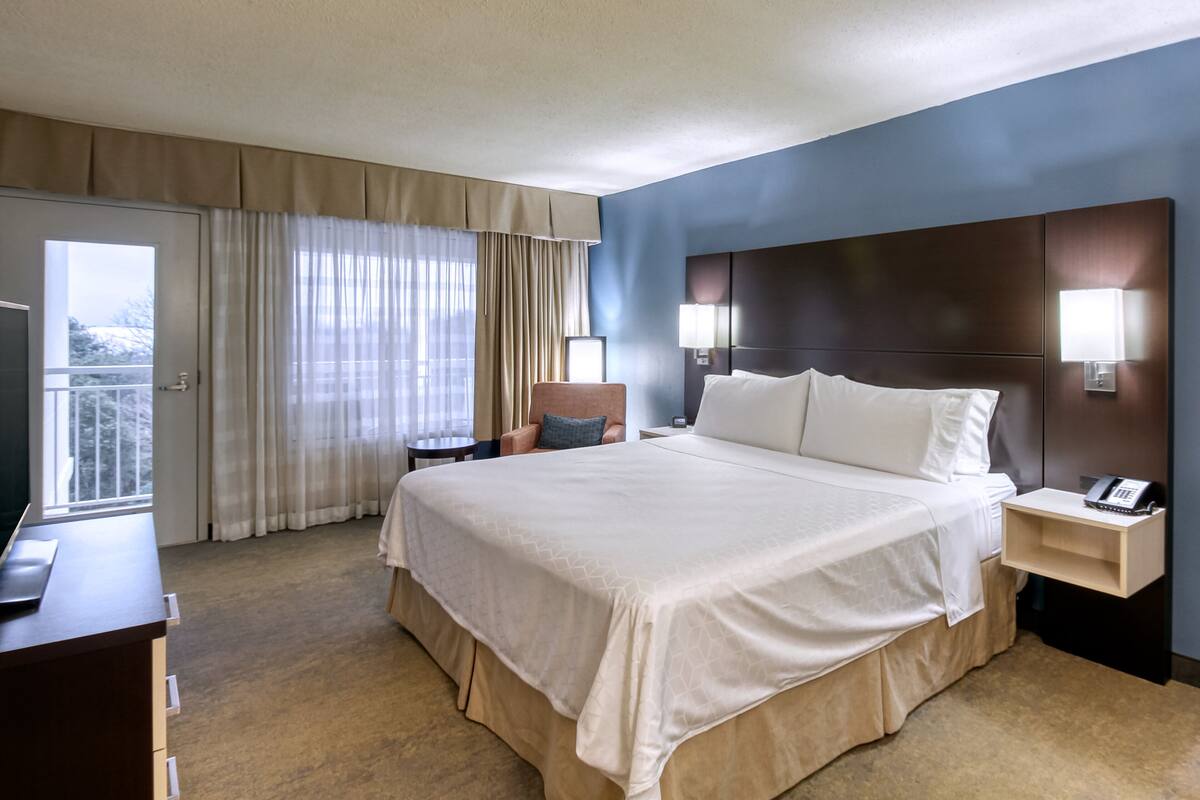 A hotel room with a large bed, a desk, a chair, a nightstand with a phone, and a view of the outdoors through the window.