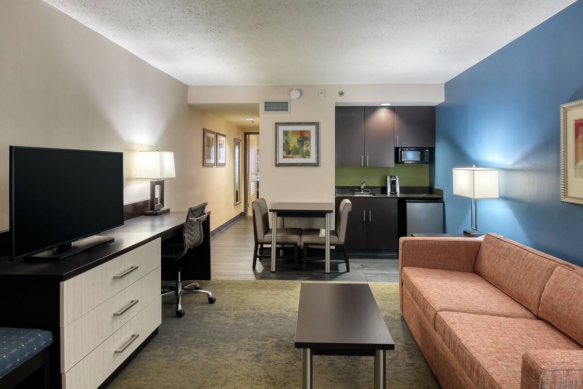 A hotel room with a TV, desk, chair, sofa, dining table, and kitchen area.