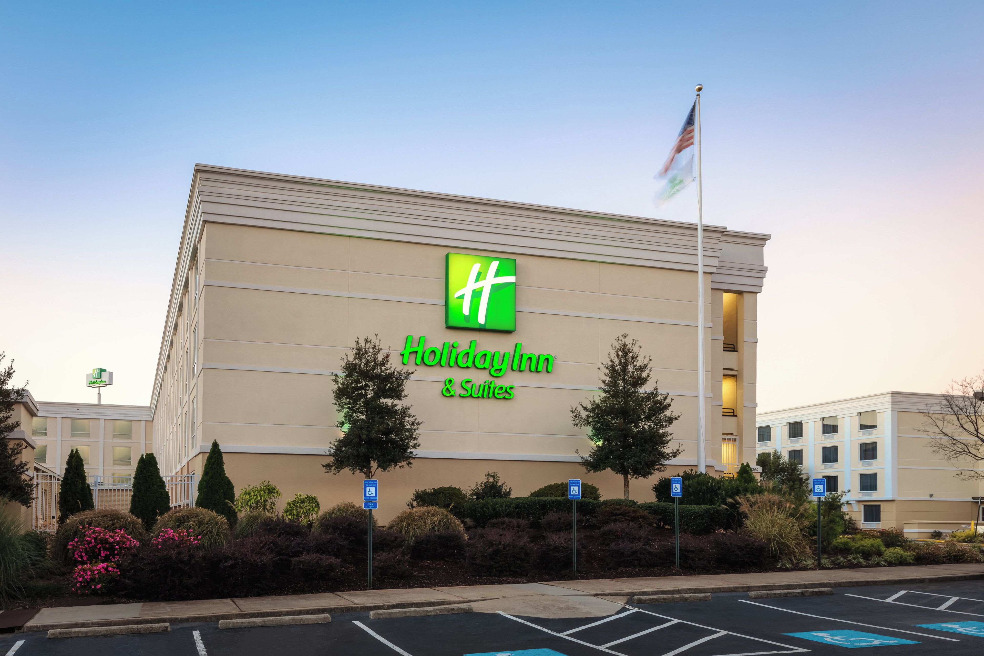 A Holiday Inn & Suites building with a prominent green logo, parking spaces, and a flag on a flagpole.