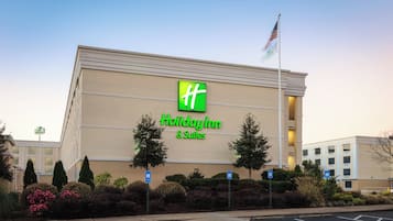A Holiday Inn & Suites building with a prominent green logo, parking spaces, and a flag on a flagpole.