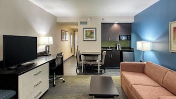 A hotel room with a television, desk, chair, sofa, kitchenette, and artwork on the walls.