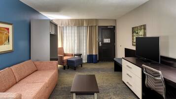 Habitación de hotel con sofá, una mesita, televisor, escritorio con cajones, una silla y ventana con cortinas.