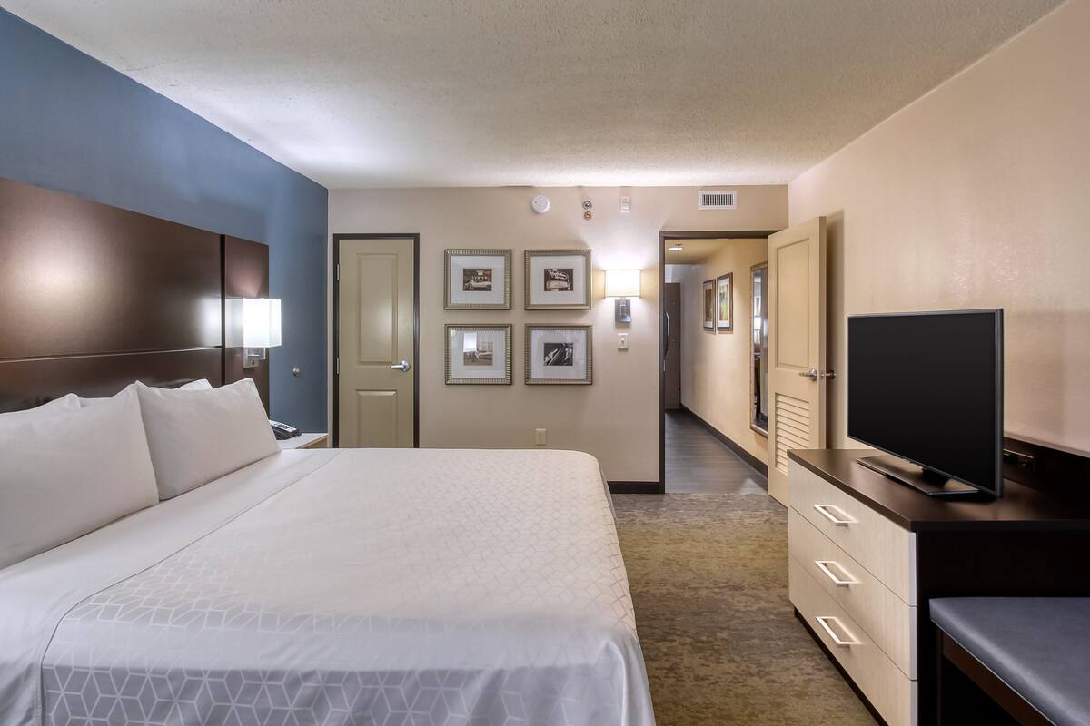 A hotel room with a large bed, a TV on a dresser, a desk with a lamp, and framed pictures on the wall.