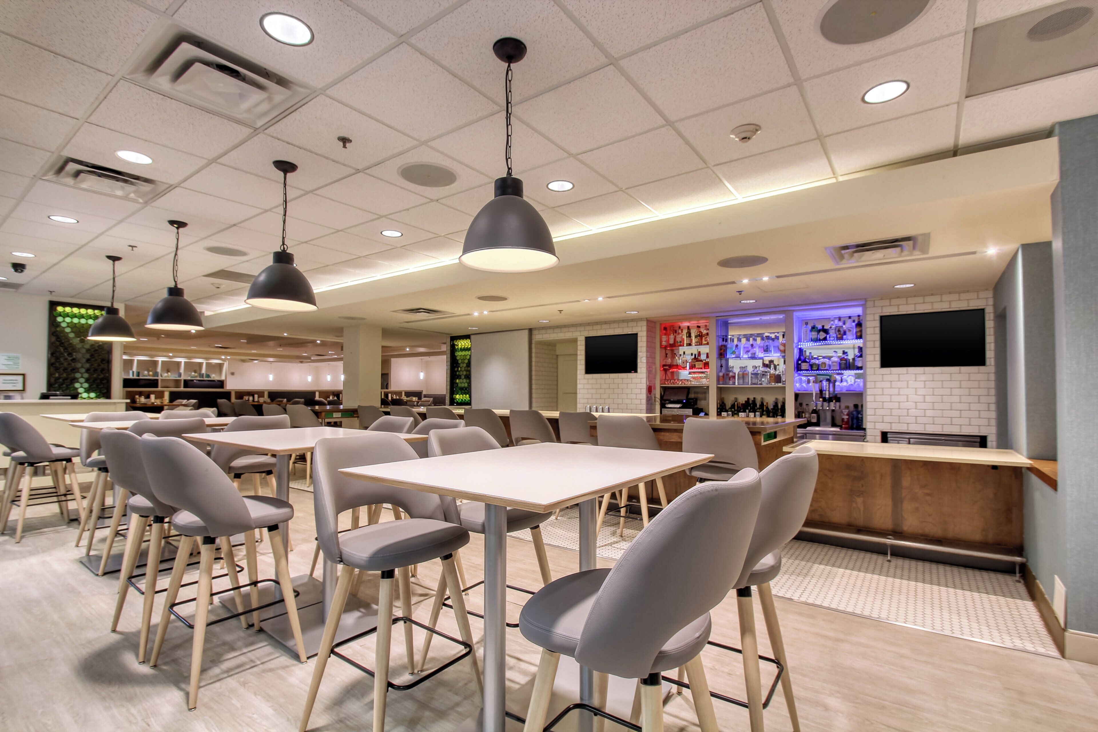 A modern restaurant interior with multiple tables and chairs, pendant lighting, a bar area, and a television screen.