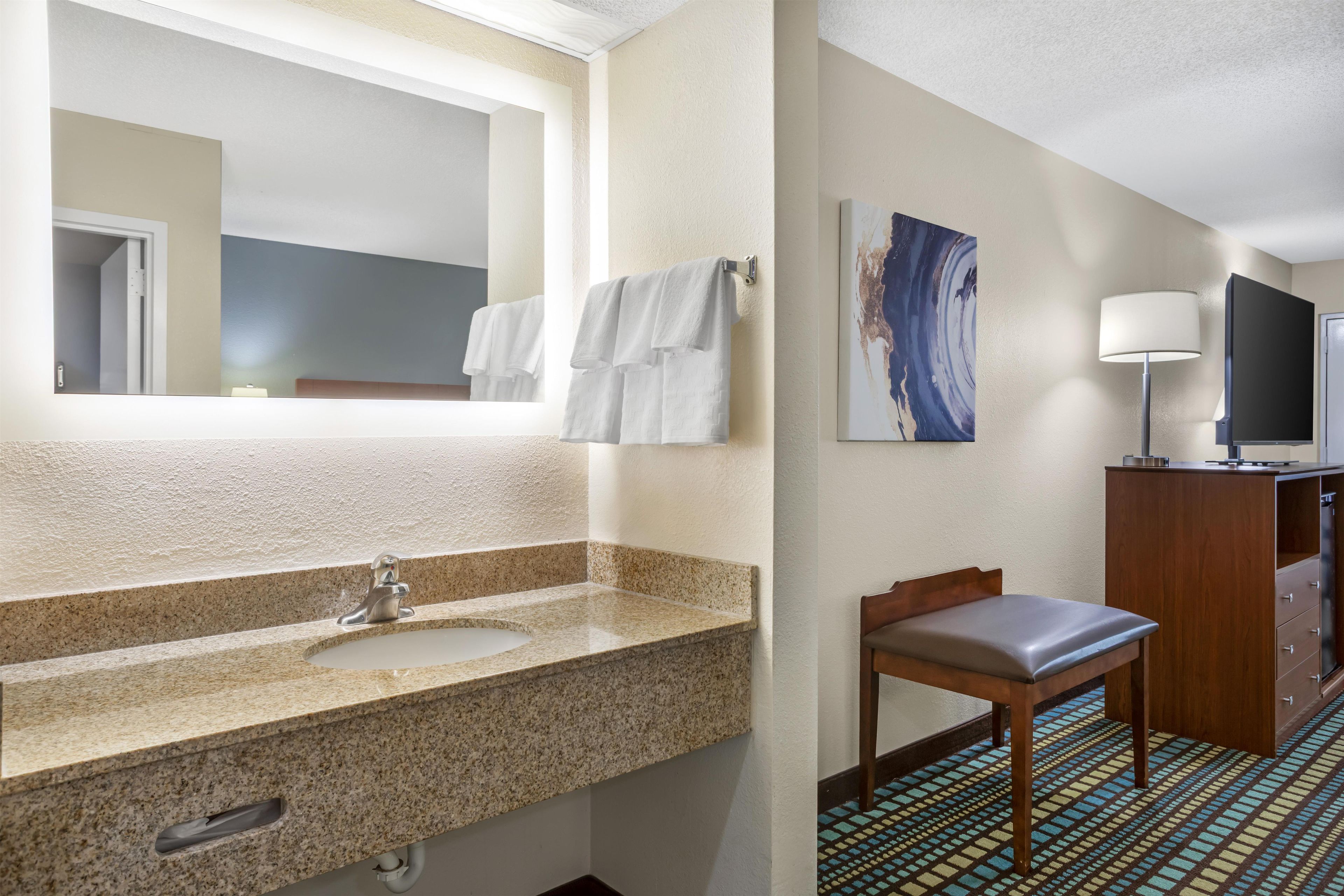 A hotel bathroom with a granite sink, a mirror, a towel rack, a wooden chair, a painting on the wall, a television, and a cabinet.