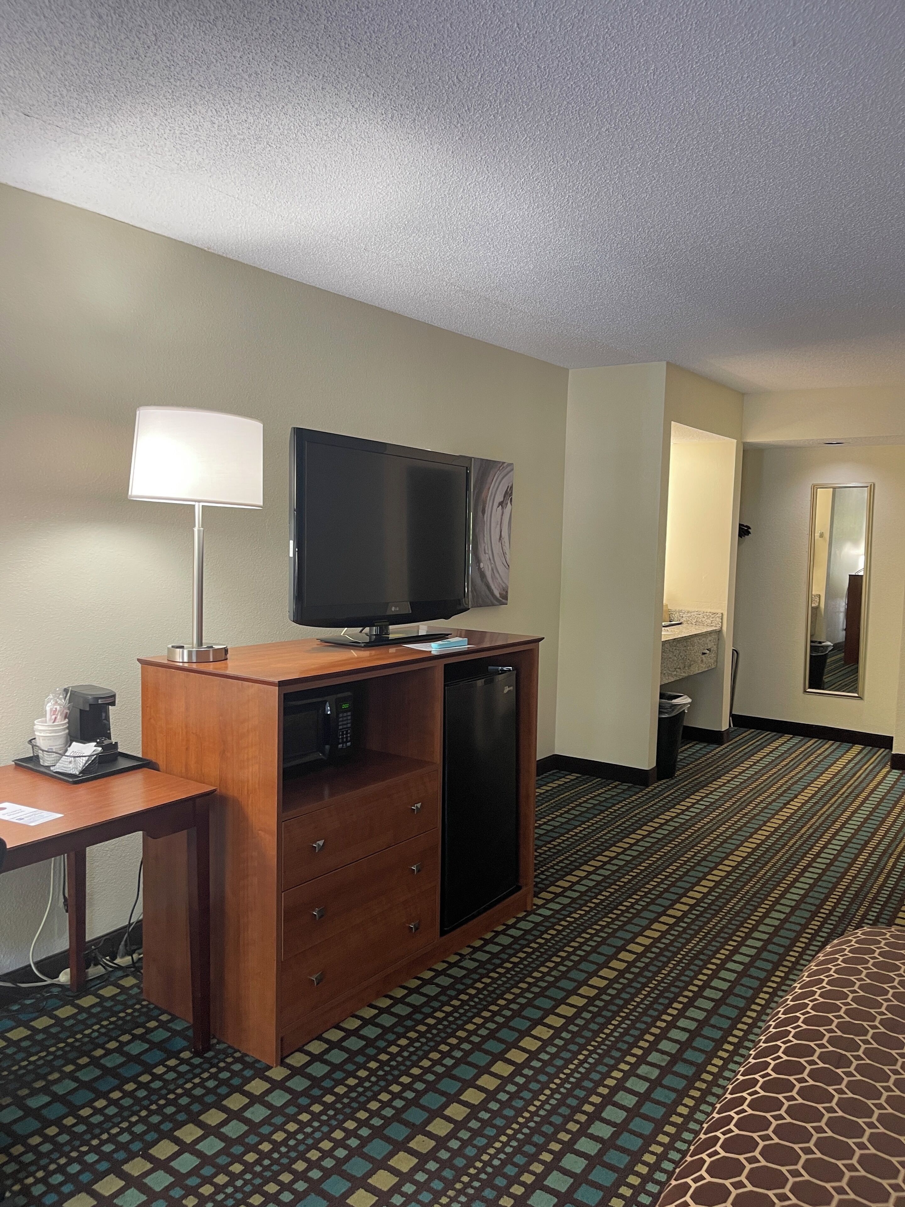 A hotel room with a wooden desk, a flat-screen TV, a coffee maker, a microwave, a refrigerator, a patterned carpet, and a mirror on the wall.